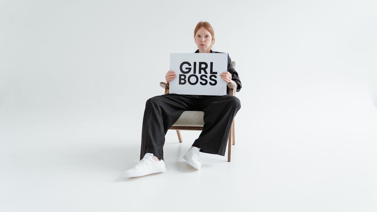 A woman in a black suit holds a 'Girl Boss' sign, exuding confidence on a plain background.